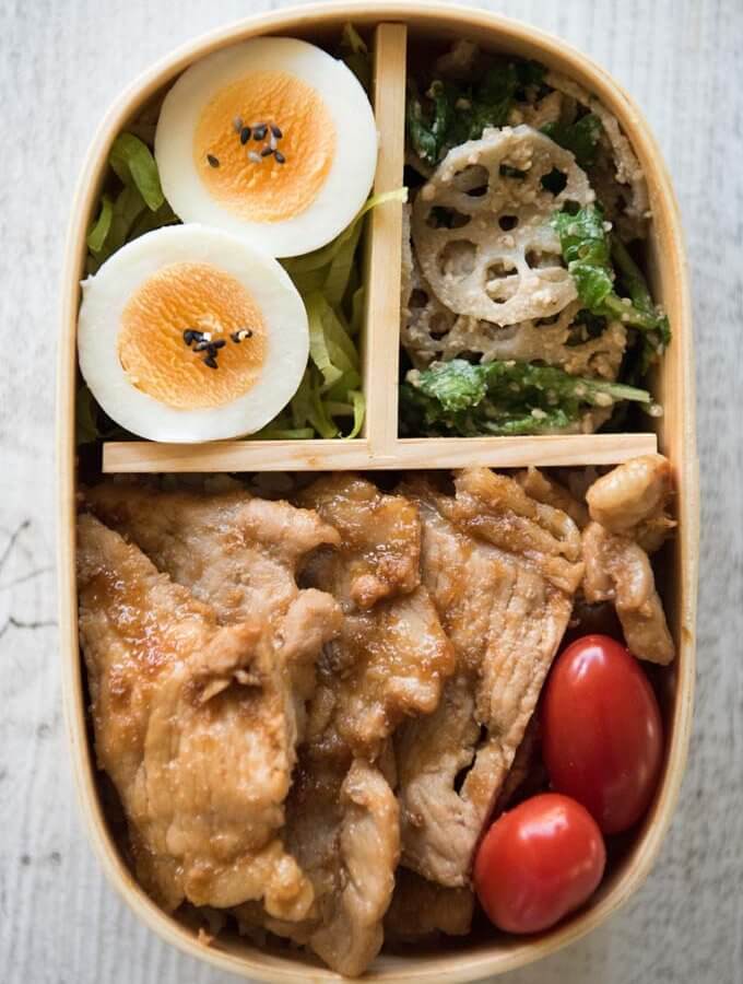 Top-down photo of Pork Shōgayaki Bento which contains Pork Shōgayaki, small tomatoes, boiled eggs on shredded lettuce and Lotus Root and Spinach salad.